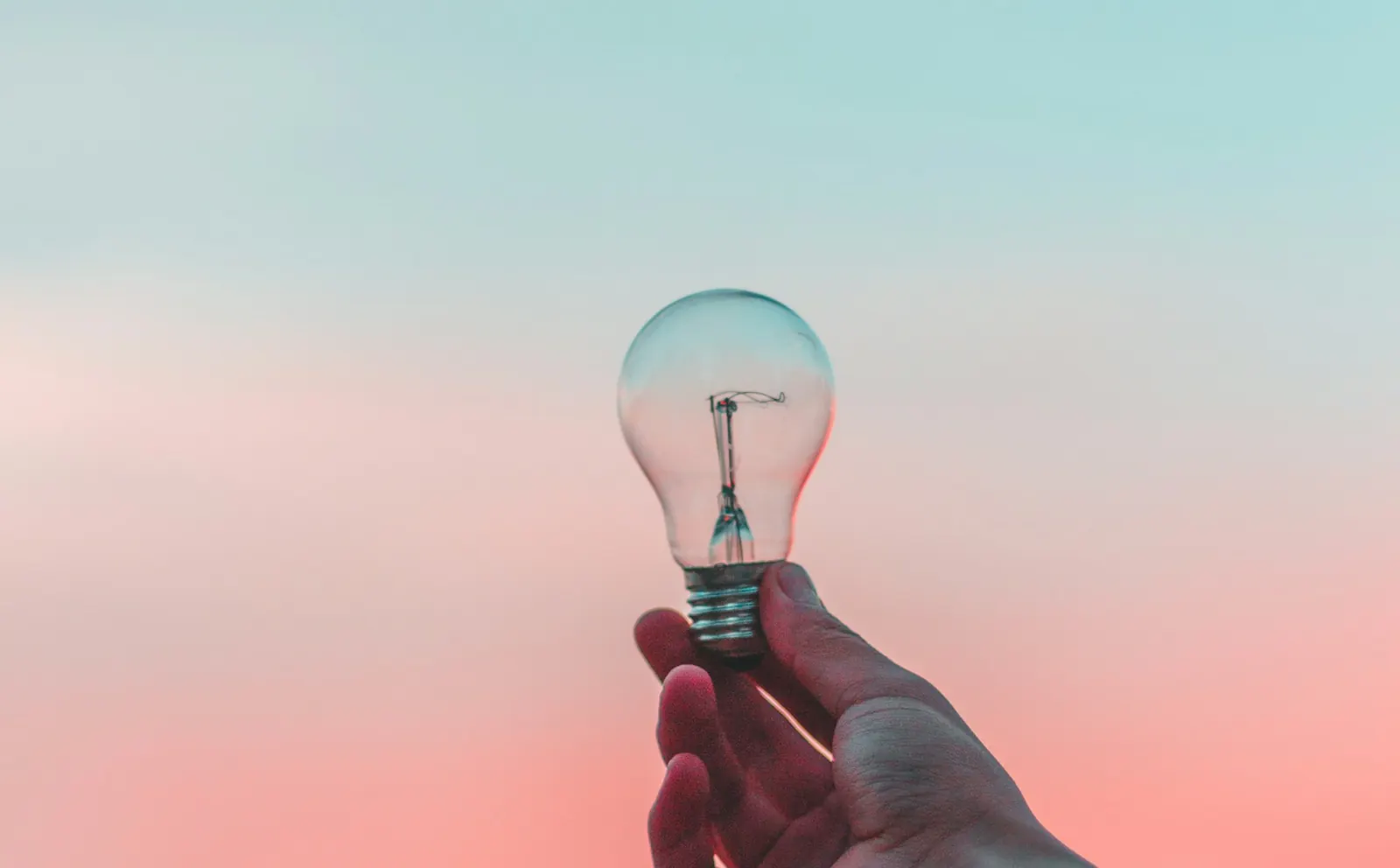 A hand holding up a light bulb against a pastel sky